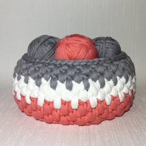 May include: A crocheted basket with a gray, white, and coral color pattern. The basket is filled with three balls of yarn, two gray and one coral.