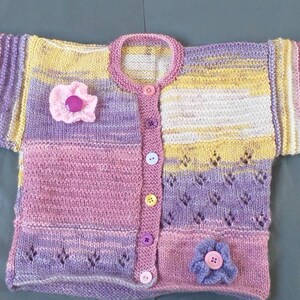 May include: A knitted baby cardigan with a striped pattern in shades of pink, purple, and yellow. The cardigan has a button closure and features two flower embellishments.
