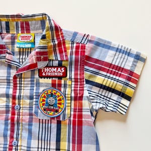 May include: A short-sleeve button-down shirt featuring a red, yellow, white, and blue plaid pattern. The shirt includes a Thomas & Friends brand tag, a rectangular logo patch, and a circular patch depicting the Thomas the Tank Engine character.