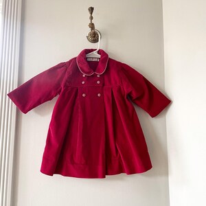 May include: A red velvet coat with a white gingham collar and pearl-like buttons. The coat has long sleeves and a flared skirt.