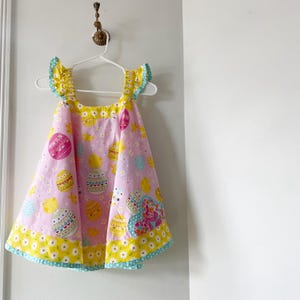 May include: A pink Easter-themed dress with a yellow and blue trim. The dress features a pattern of Easter eggs and chicks. The shoulders have ruffled sleeves with yellow and blue accents. The dress is hanging on a white hanger.