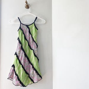 May include: A sleeveless dress with a floral daisy pattern in pink and white, alternating with green panels. The dress has black trim with small white polka dots and thin navy blue straps. The dress is hanging on a white hanger.
