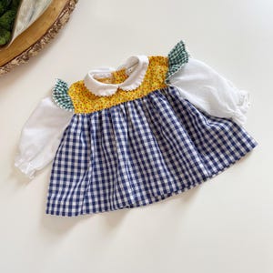 Puede incluir: Un vestido de bebé de cuadros vichy blanco, azul y amarillo con cuello blanco y mangas con volantes verdes y blancos.