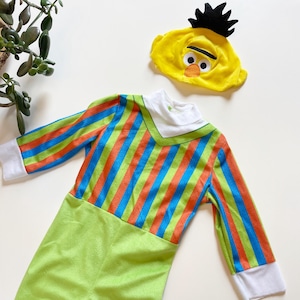 Puede incluir: Un disfraz infantil con un sombrero de personaje amarillo con pelo negro y un mono a rayas a juego. El mono tiene rayas verdes, naranjas y azules en la parte superior y una parte inferior verde lisa. El disfraz tiene puños blancos y un cuello blanco.