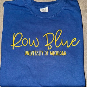 May include: A blue t-shirt with the words "Row Blue" in yellow script and "University of Michigan" in yellow text below.