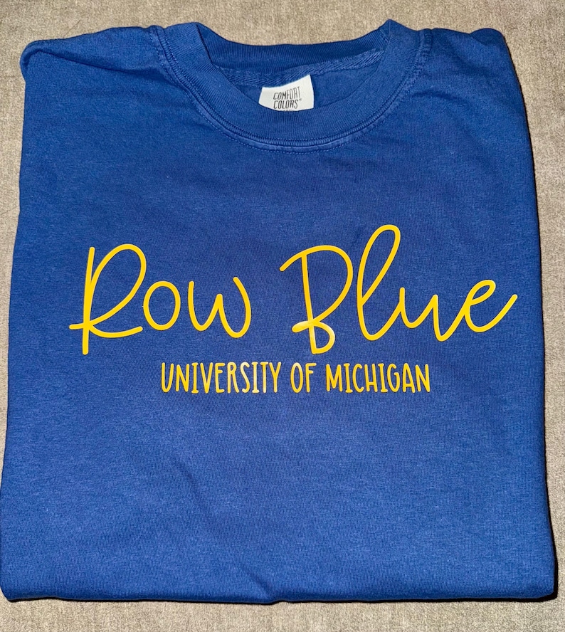May include: A blue t-shirt with the words "Row Blue" in yellow script and "University of Michigan" in yellow text below.