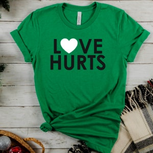 Może przedstawiać: Zielona koszulka z białym sercem i napisem "LOVE HURTS" w kolorze czarnym.