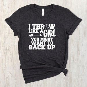 May include: A dark grey t-shirt with white text that reads "I throw like a girl you might want to back up". A white baseball is included in the text.