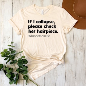 May include: Cream-colored t-shirt with the text "If I collapse, please check her hairpiece." and the hashtag "#dancemomlife" in black. The shirt is knotted at the bottom. A brown hat and greenery are in the background.