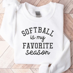 以下が含まれることがあります： 黒文字で「Softball is my favorite season」とプリントされた白いスウェットシャツです。
