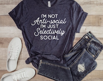 May include: A navy blue t-shirt with white text that reads "I'm not anti-social I'm just selectively social". The shirt is laid out on a wooden surface with a pair of white trainers and blue jeans.