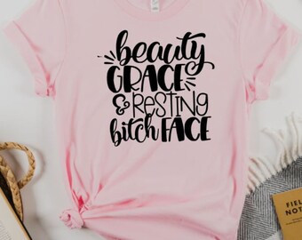 May include: Pink t-shirt with black text that reads "beauty grace & resting bitch face"