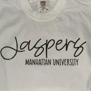 May include: White t-shirt with black glitter lettering that reads "Jaspers Manhattan University".
