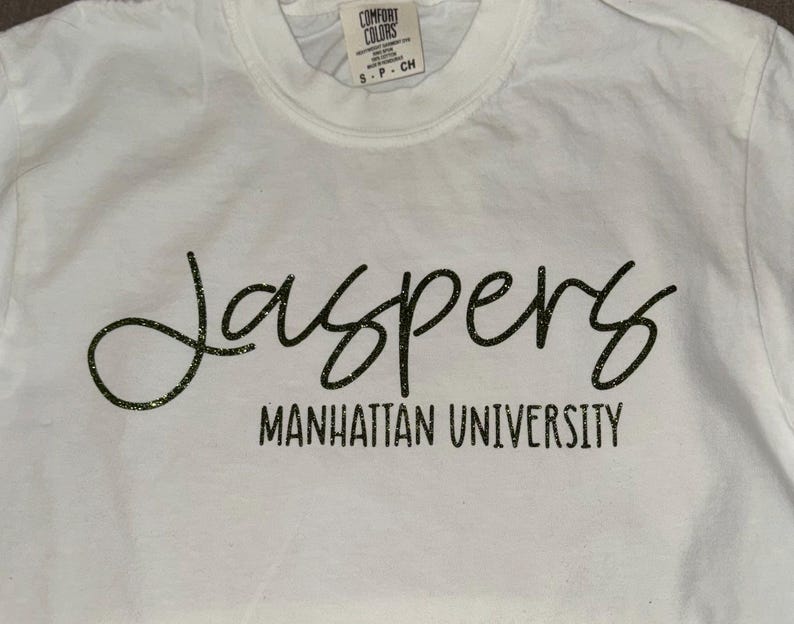 May include: White t-shirt with black glitter lettering that reads "Jaspers Manhattan University".