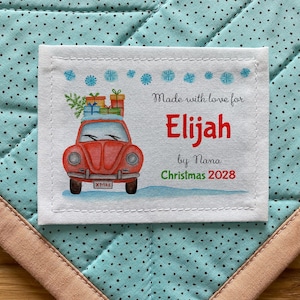 Peut inclure: Une étiquette personnalisée avec une voiture rouge chargée de cadeaux, des flocons de neige et le texte "Made with love for Elijah by Nana Christmas 2028." L'étiquette est sur un tissu bleu à pois avec une bordure rose.
