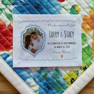 Pode incluir: Um rótulo de tecido branco com costuras azuis e uma foto de um casal. O rótulo diz "Made especially for LARRY & STACY In celebration of their marriage on March 11, 2026 Love, Mom"