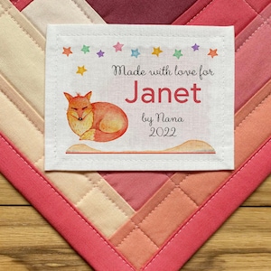 May include: A white fabric label with a sleeping fox illustration, colorful stars, and the text "Made with love for Janet by Nana 2022".