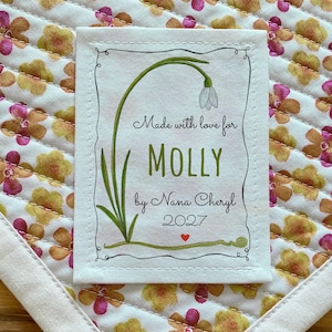 Op de afbeelding: Een wit stoffen label met een zwarte rand en een groen bloemmotief. Het label luidt "Made with love for MOLLY by Nana Cheryl 2027" met een klein rood hart.