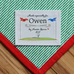 May include: A white fabric label with green and white striped fabric behind it. The label says "Made especially for Owen by Auntie Laurie 2027" with a red and blue flag, golf clubs, and a golf ball on a green hill.