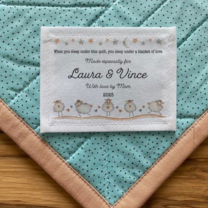 Peut inclure: Une étiquette de couette personnalisée avec le texte "Made especially for Laura & Vince" et "With love by Mom 2025". L'étiquette présente une illustration de moutons et est cousue sur une couette turquoise à motifs de pois et bordure beige.