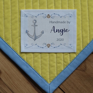 May include: A handmade quilt label with an anchor design, framed by decorative elements. The label reads "Handmade by Angie 2020." The quilt has a yellow quilted center and a blue border.