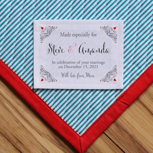 Op de afbeelding: Een wit stoffen label met zwarte tekst en rode harten. Het label luidt "Made especially for Steve & Amanda In celebration of your marriage on December 15, 2021 With love from Mom".