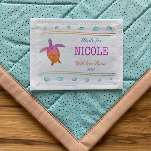 May include: A white fabric label with a hand-drawn illustration of a sea turtle with orange, pink, and yellow colors. The label says "Made for NICOLE With love, Nana 2021".