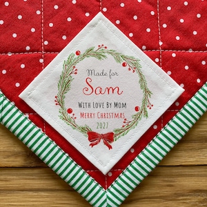 Può includere: Un runner natalizio trapuntato con un motivo a pois rossi e un bordo a righe verdi e bianche. Una toppa quadrata bianca al centro recita "Made for Sam, With Love by Mom, Merry Christmas 2027."