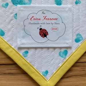 以下が含まれることがあります： 雲の形をした縁取りのある白い布のラベルに、「Erica Ferroneへ  ナナから愛情込めて手作り 2021」と書いてあります。ラベルの中央には、黒い斑点のある赤いテントウムシがいます。