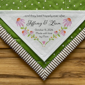 May include: A white triangular fabric label with the text "...and they lived happily ever after. Tiffany & Liam October 15, 2026 Made with love by Mom" and a floral design.