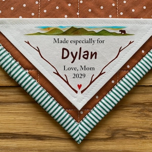 May include: A personalized, triangular fabric label with the text "Made especially for Dylan, Love, Mom 2029." The label features a mountain and bear illustration. The label is bordered by green and white striped fabric and brown fabric with white polka dots.