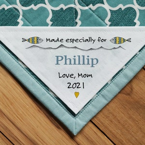 Pode incluir: Etiqueta triangular de tecido branco com ilustrações de peixes azuis e amarelos e o texto "Made especially for Phillip Love, Mom 2021" com um pequeno coração amarelo.