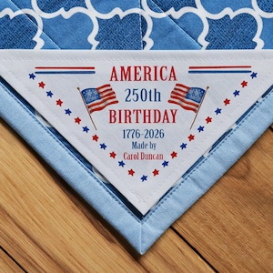 Pode incluir: Um canto de colcha patriótica com um rótulo celebrando o 250º aniversário da América. O rótulo apresenta o texto "AMERICA 250th BIRTHDAY 1776-2026" com bandeiras americanas e estrelas vermelhas e azuis. A colcha tem um padrão azul e branco.