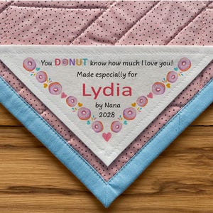 May include: A white triangular fabric label with pink polka dots and a blue border. The label has a donut-themed design with the text "You DONUT know how much I love you! Made especially for Lydia by Nana 2028".