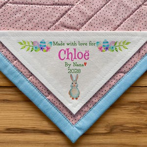 May include: A personalized baby blanket with a white triangular label. The label reads "Made with love for Chloe" with a bunny illustration. The blanket has a pink polka dot pattern, blue trim, and is set on a wooden surface.