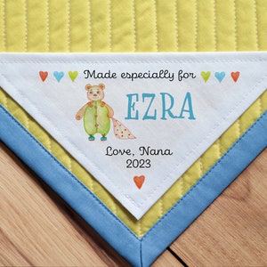May include: White triangular fabric label with a hand-drawn illustration of a teddy bear wearing a green onesie and holding a pink blanket. The label reads "Made especially for EZRA Love, Nana 2023" with colorful hearts surrounding the text.