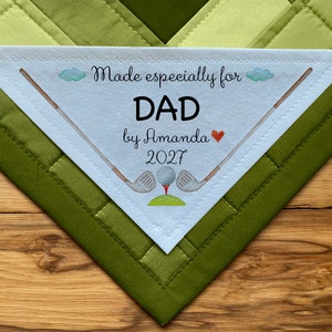 Könnte beinhalten: Weißer dreieckiger Stoff-Patch mit einer gestickten Umrandung. Es steht "Made especially for DAD by Amanda 2027" mit einem roten Herzen. Es gibt zwei Golfschläger und einen Golfball auf einem grünen Fleck.