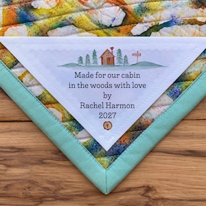 May include: A white triangular fabric label with a hand-drawn illustration of a cabin in the woods. The text reads "Made for our cabin in the woods with love by Rachel Harmon 2027".