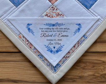 Könnte beinhalten: Ein weißes Stoffquadrat mit blauer Blumenstickerei und dem Text "Your wedding day will come and go, but may your love forever grow. Robert & Emma October 21, 2020 Love, Mom"