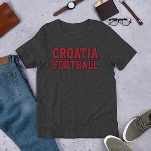 May include: Dark heather grey t-shirt with red text that says "CROATIA FOOTBALL" in a collegiate font.