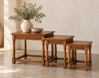 May include: A set of three graduated wooden nesting tables with square tops and turned legs. The largest table holds a textured cream-colored ceramic vase filled with green leafy branches and a small matching ceramic bowl.