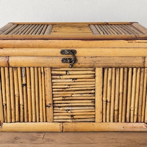 May include: A rectangular storage box constructed from light brown bamboo stalks. The exterior features vertical and horizontal bamboo patterns with a dark metal latch centered on the front panel to secure the hinged lid.