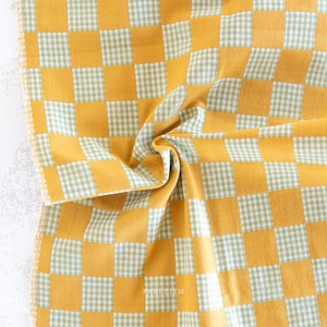 May include: A close-up shot of a fabric featuring a yellow and green checkered design. The fabric is folded, revealing its texture and pattern. The squares alternate between solid yellow and a green and white gingham.
