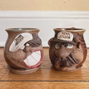 May include: Two brown ceramic mugs with expressive, cartoonish faces. One mug has a white hat with "HOT STUFF" text, and the other has a similar hat. The mugs have a rustic, handmade appearance, with visible textures and details.