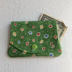 May include: A small green fabric coin purse with a white polka dot pattern and colorful teapots. The purse has a green button closure and a dollar bill is sticking out of the top.
