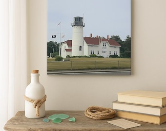 May include: A white ceramic bottle with a cork stopper and a rope tied around it sits on a wooden table. A canvas print of a white lighthouse with a red roof and flags hangs in the background. Sea glass and books are also on the table.