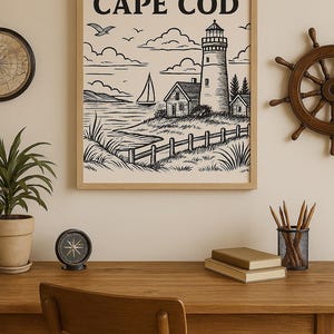 May include: Framed black and white Cape Cod art print featuring a lighthouse, sailboat, and coastal landscape. The print is displayed on a wooden desk with a compass, pencils, and a ship's wheel.