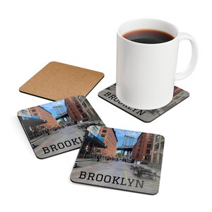May include: A white ceramic mug filled with coffee sits on a coaster with a Brooklyn street scene. Two additional coasters with the word "BROOKLYN" and a Brooklyn street scene are also visible, along with a plain cork coaster.