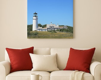 May include: A framed canvas print of a coastal scene featuring a white lighthouse with a black top, a gray building, and a blue sky. The print hangs above a white sofa with red and cream-colored pillows. A wooden side table holds a white mug.