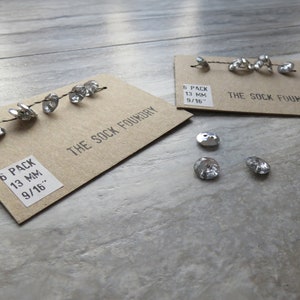 Puede incluir: Seis botones plateados con superficie texturizada, dispuestos en una tarjeta marrón con el texto "6 PACK 13 MM 9/16" y "THE SOCK FOUNDRY".